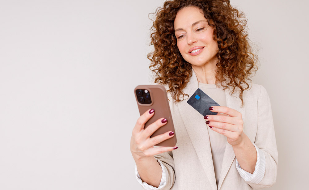 A woman holds a smartphone and payment card while standing against a plain light background.
