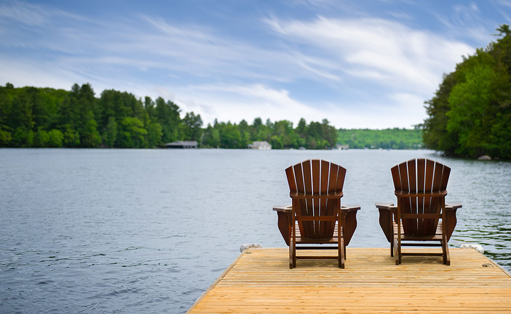 Two wooden chairs sit on a dock overlooking a calm lake surrounded by trees.