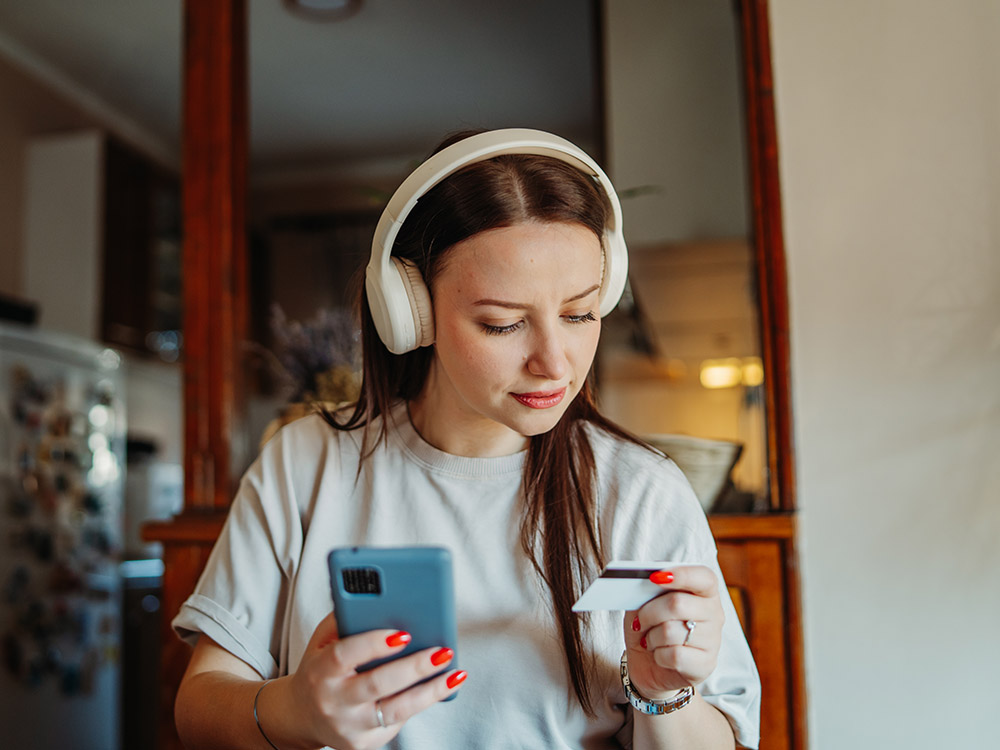 A young woman wearing headphones looks at a card while using a smartphone.