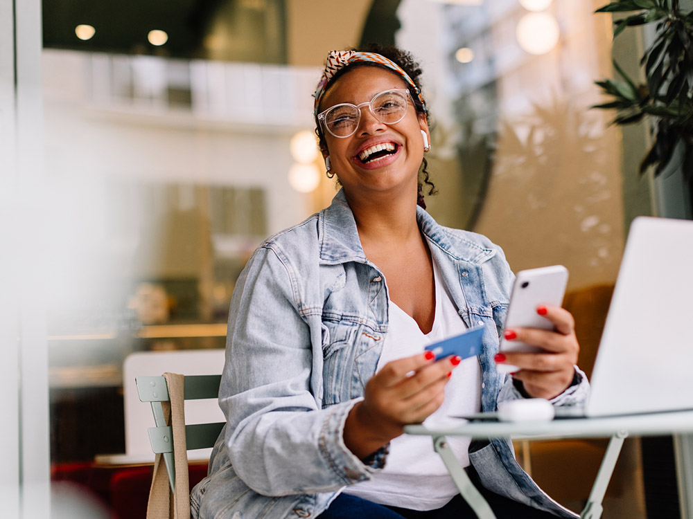 750_0001_iStock-1956924237 Smiling woman seated at a cafe holds a smartphone and payment card near a laptop.