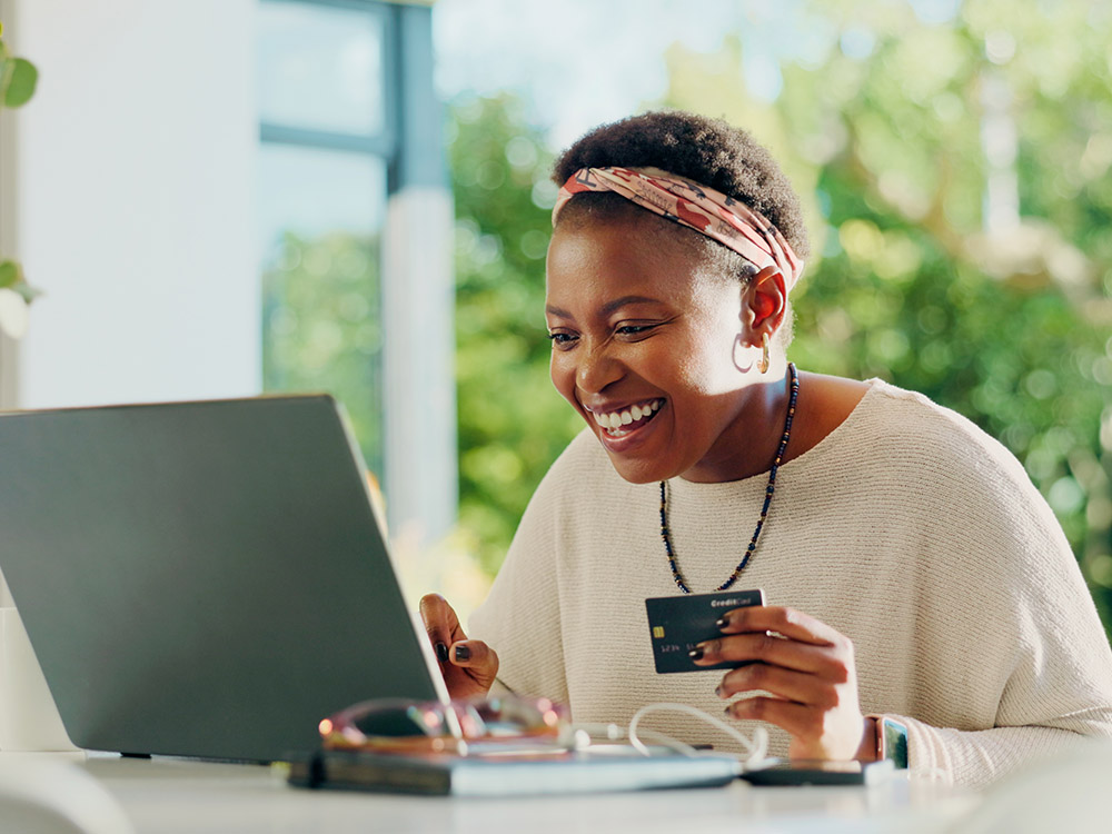 750_0001_iStock-2230456722 A woman smiles while holding a payment card and working on a laptop.