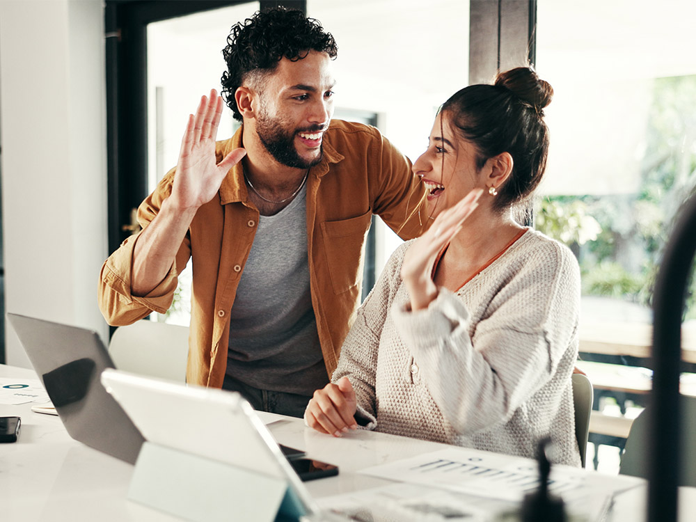 Two coworkers smile and wave during a video call from a desk with digital devices.