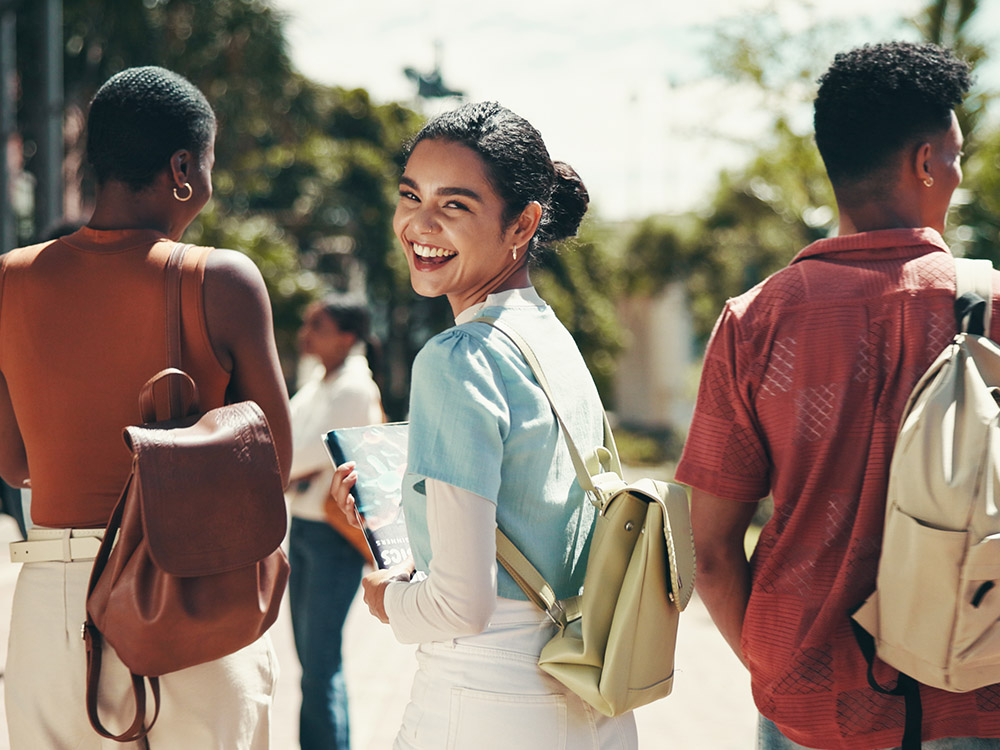 750_0007_iStock-2199077254 Smiling students walk outdoors on campus, carrying notebooks and backpacks in bright daylight.