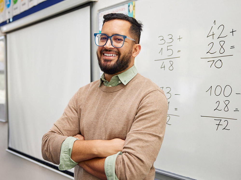 750_0008_iStock-2160473960 A smiling teacher stands in front of a classroom whiteboard with math problems.