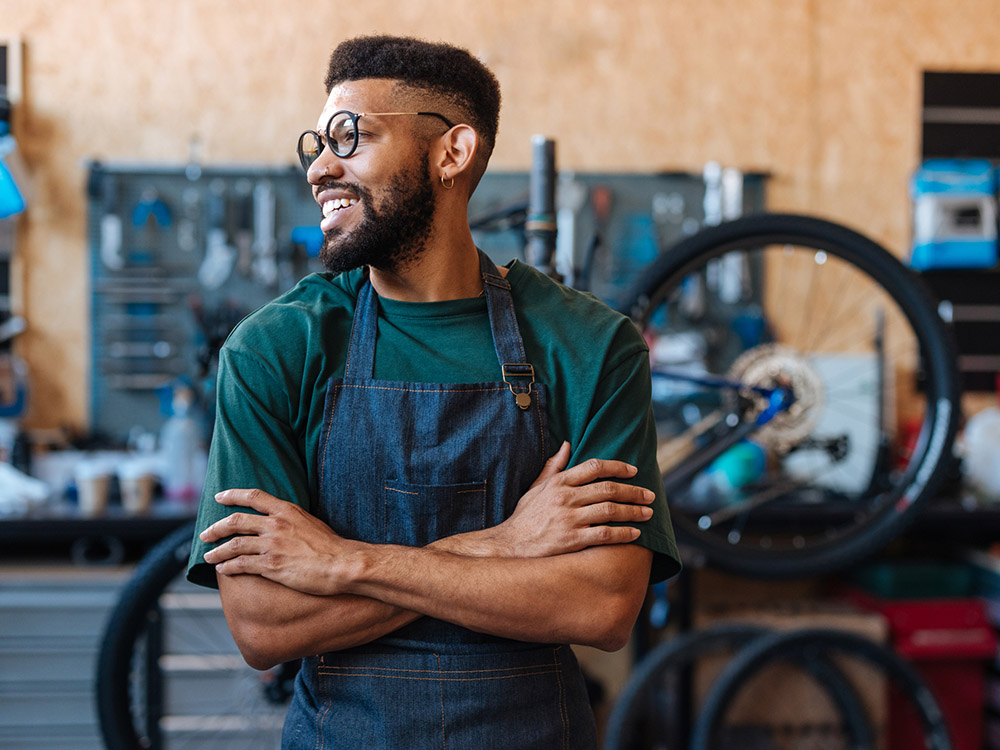 750__0001_iStock-2232668645 A bicycle shop worker smiles with arms crossed in front of bikes and repair tools.