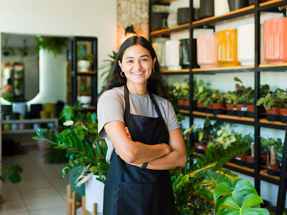 Smiling plant shop worker stands with arms crossed in front of shelves filled with potted plants