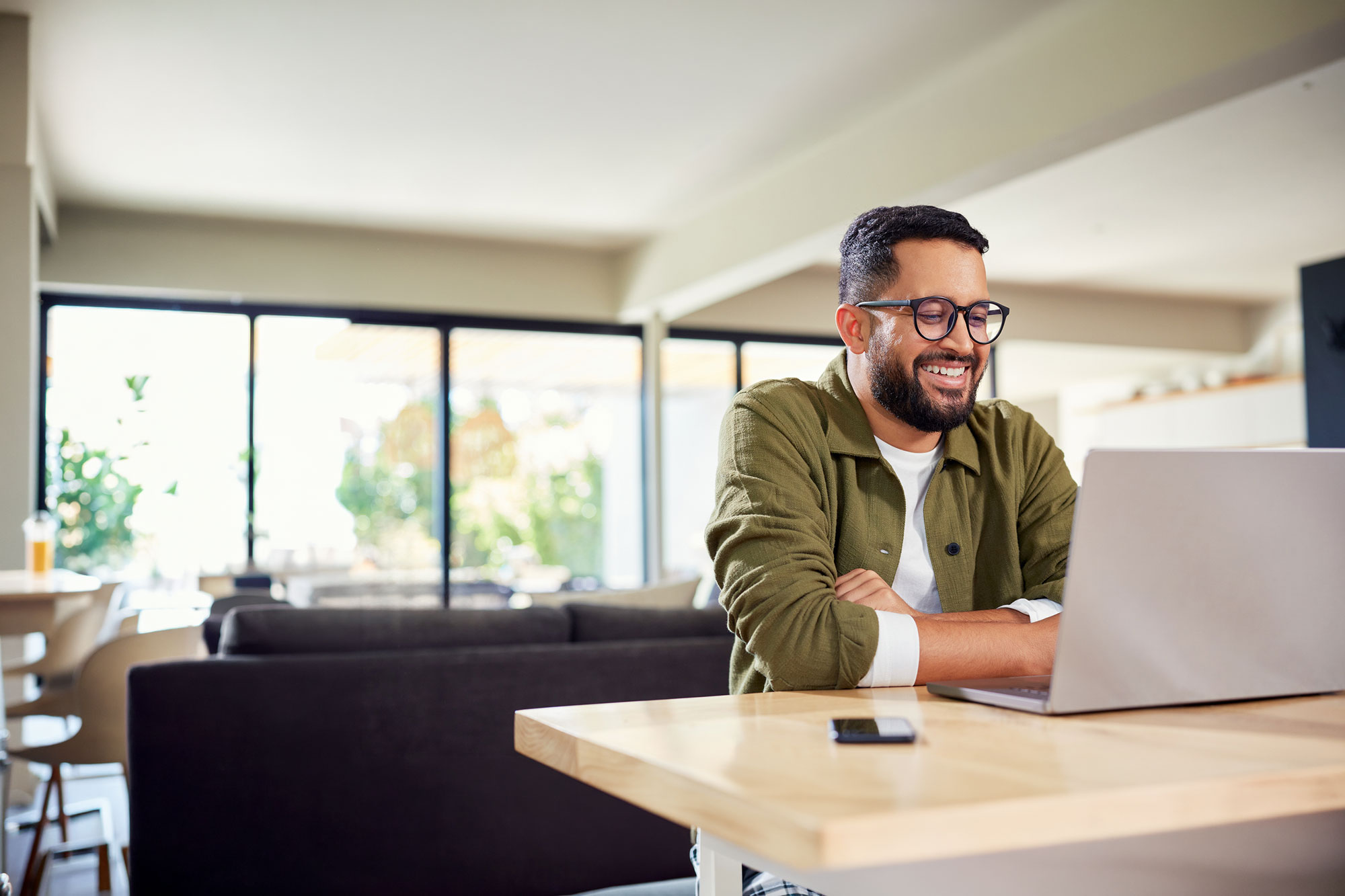 Smiling man with glasses works on a laptop at a table in a bright home.