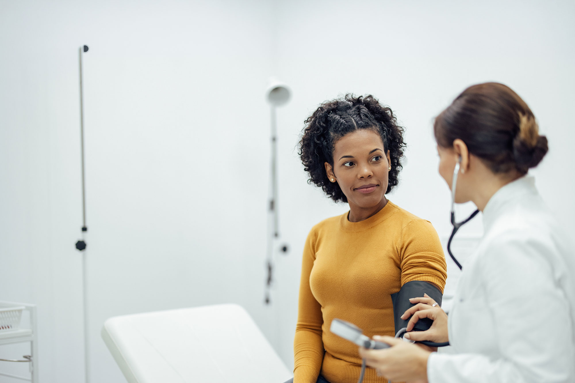 A patient sits in an exam room while a healthcare provider listens to her arm with a stethoscope and discusses her care.