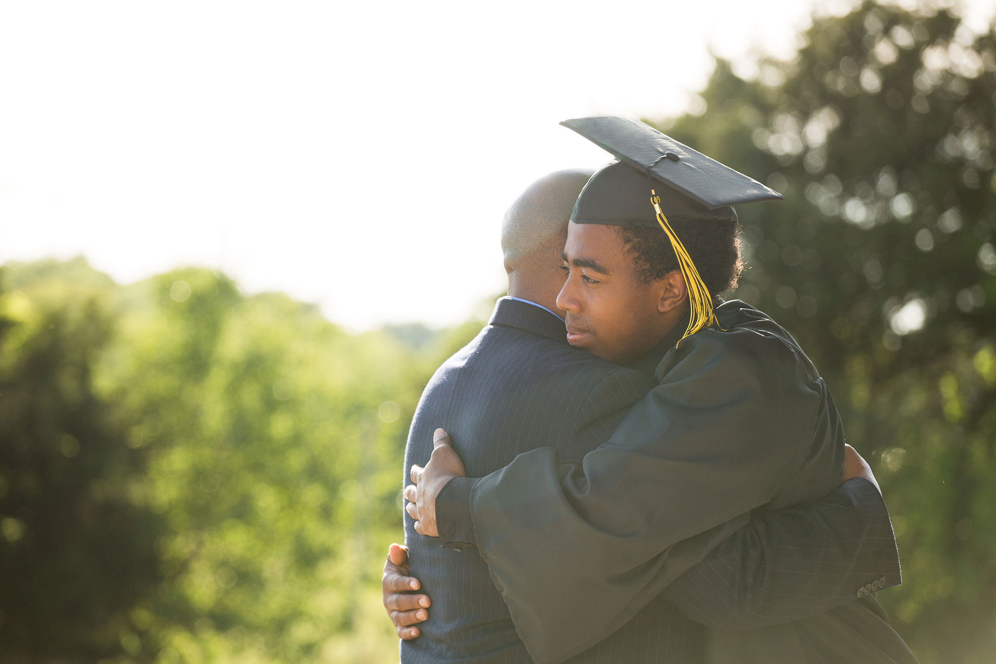 Graduate in cap and gown embraces an older family member outdoors in warm sunlight.