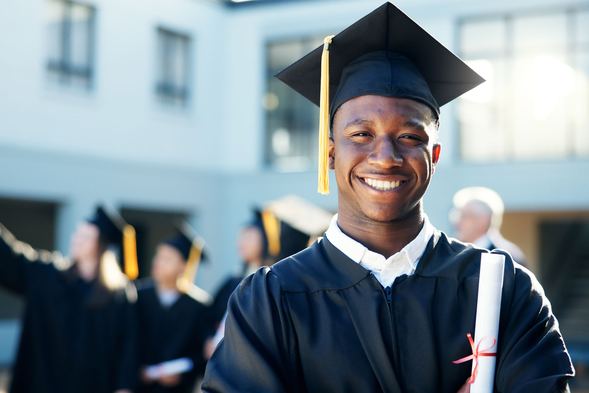 A smiling graduate in a cap and gown holds a diploma while other graduates stand blurred in the background.