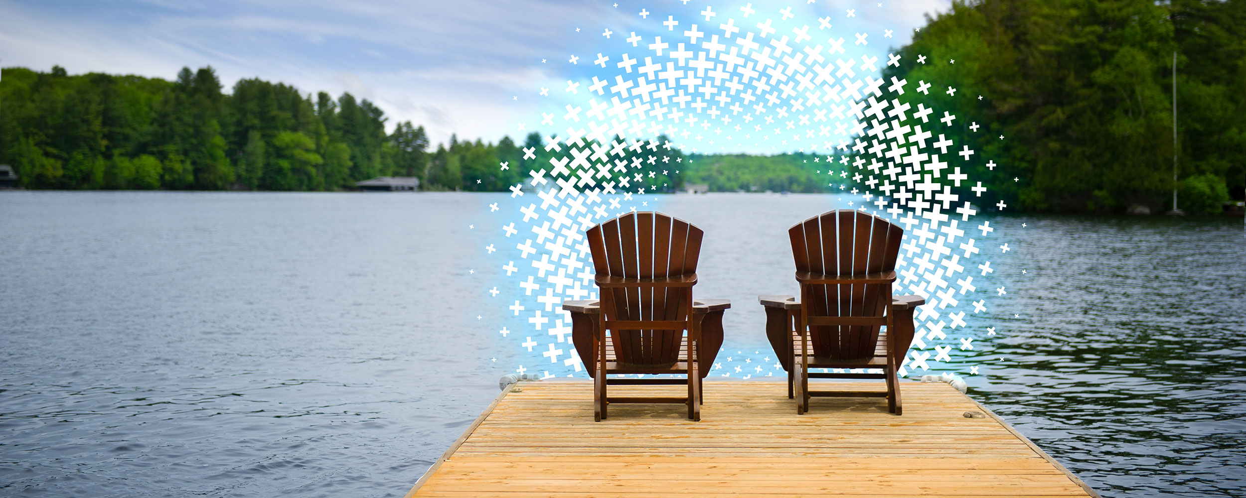 Two Adirondack chairs on a wooden dock overlooking a calm lake surrounded by trees.