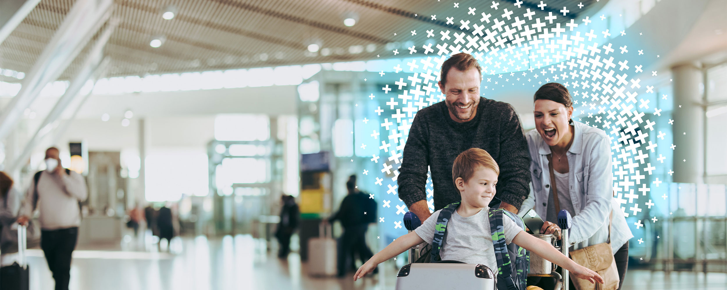 Family traveling through an airport with their young child riding on a luggage cart.