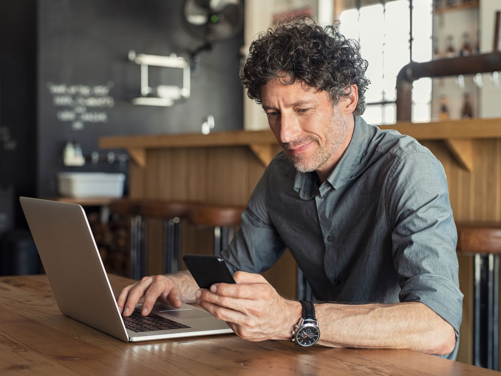 Man using a smartphone and laptop while seated at a table.