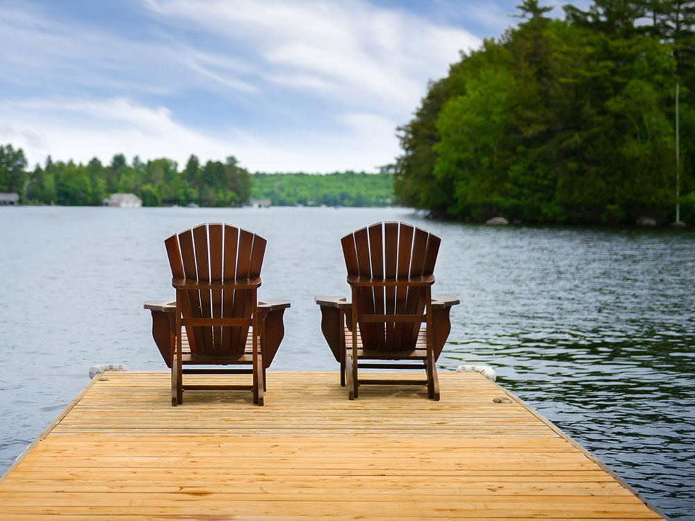 Two Adirondack chairs on a wooden dock overlooking a calm lake surrounded by trees.