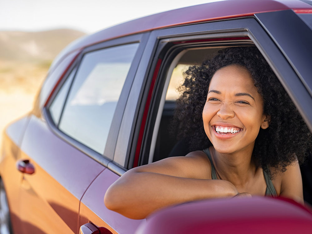 A smiling woman leans out of a car window with her arm resting on the door during a sunny drive.