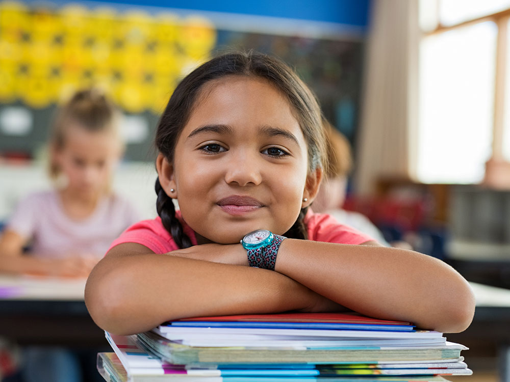 Student resting her arms on a stack of books and smiling in a classroom.